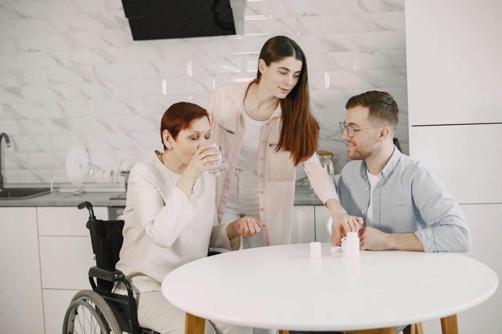 A woman in a wheelchair drinks water at a kitchen table. A younger woman and man assist her, creating a supportive and caring atmosphere.