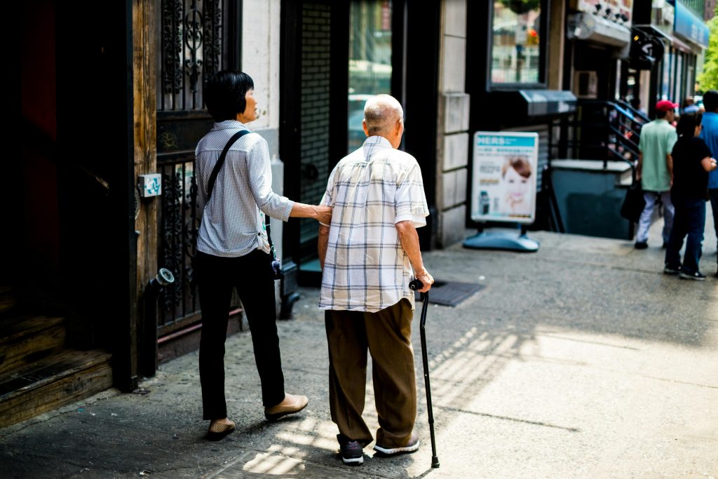 A person supports an elderly man with a cane, walking down a sunlit city street. They appear caring and attentive, with shops and people in the background.